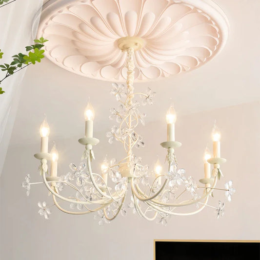 View of a white chandelier with clear crystal flower accents and candle-shaped lights against a white ceiling.