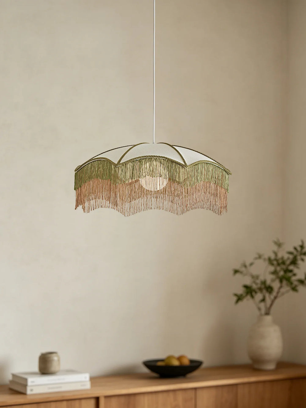 A white fabric umbrella pendant light with layered green and pink fringe hanging over a wooden cabinet.