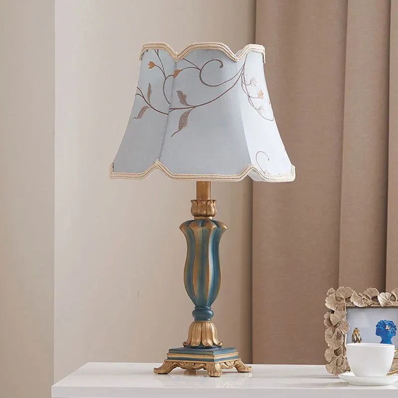 Ornate table lamp with a teal and gold resin base and a light blue fabric lampshade featuring a floral pattern, scalloped edges, and gold trim.