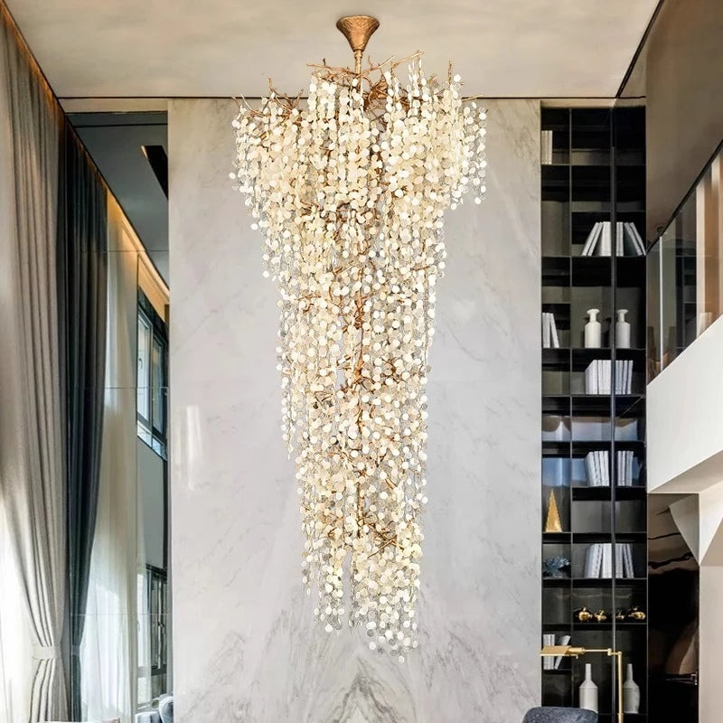 A tall, multi-tiered crystal chandelier with a gold frame illuminates a spacious living area with marble walls and a tall bookshelf.