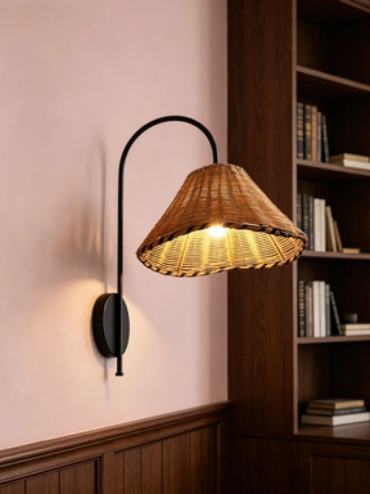 Black metal arm wall sconce with a woven rattan cone shade against a pink wall, with a bookshelf in the background.