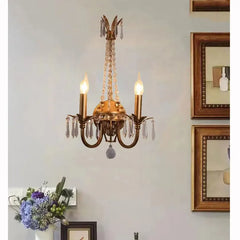 A gold crystal wall sconce with two candle-style lights mounted on a light blue wall, with framed artwork and a vase of flowers visible nearby.