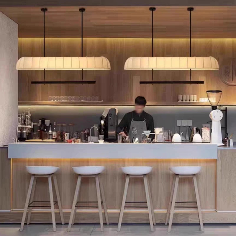 Two long, cream-colored fabric pendant lights with warm illumination, hanging above a wooden bar counter with white stools.