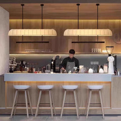 Two long, cream-colored fabric pendant lights with warm illumination, hanging above a wooden bar counter with white stools.