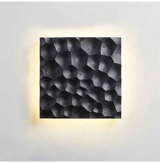 A square wall sconce with a textured, honeycomb-like pattern in a black stone finish, emitting a warm yellow glow from behind.
