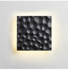 A square wall sconce with a textured, honeycomb-like pattern in a black stone finish, emitting a warm yellow glow from behind.