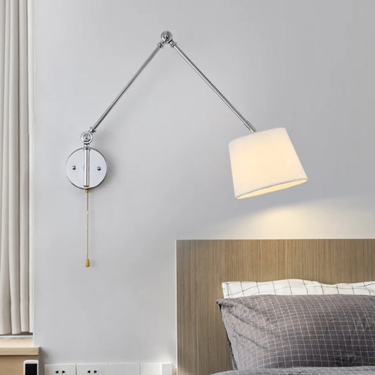 Chrome swing arm wall sconce with a white fabric drum shade extended over a bed.