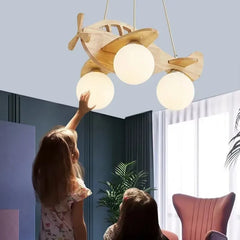 Two children reaching up towards a wooden airplane pendant light with milk white glass shades in a brightly lit room.