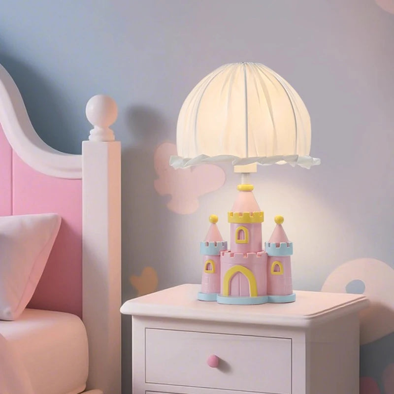 Close-up of a pink castle table lamp with a pleated white fabric shade on a white bedside table, next to a pink headboard.