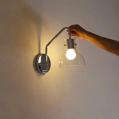 A hand holding the brushed nickel wall sconce with clear glass dome, showing the light bulb inside, mounted on a white wall.