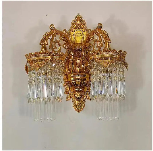 A golden wall sconce with two tiers of crystal pendants, mounted on a white wall. The fixture has ornate gold detailing.