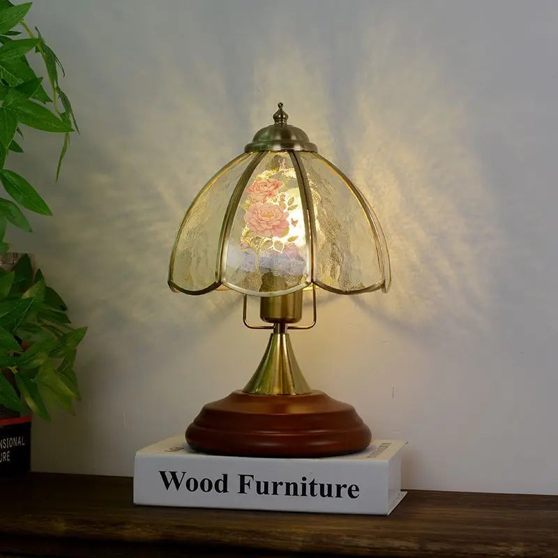 A brass table lamp with a glass shade displaying a rose floral motif, casting a warm glow on a wooden surface.