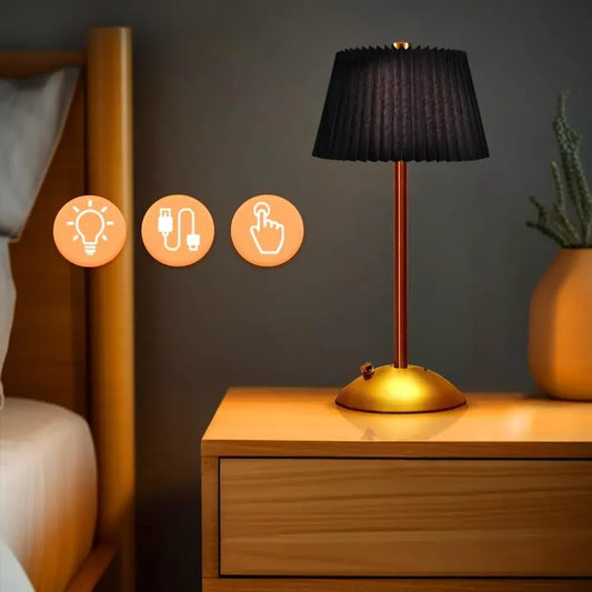 A black pleated fabric shade table lamp with a gold base and stem, placed on a wooden nightstand next to a bed.