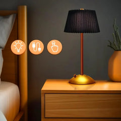 A black pleated fabric shade table lamp with a gold base and stem, placed on a wooden nightstand next to a bed.