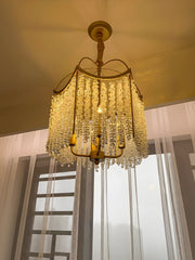 A gold crystal chandelier with cascading droplets hanging in front of a window with sheer white curtains.