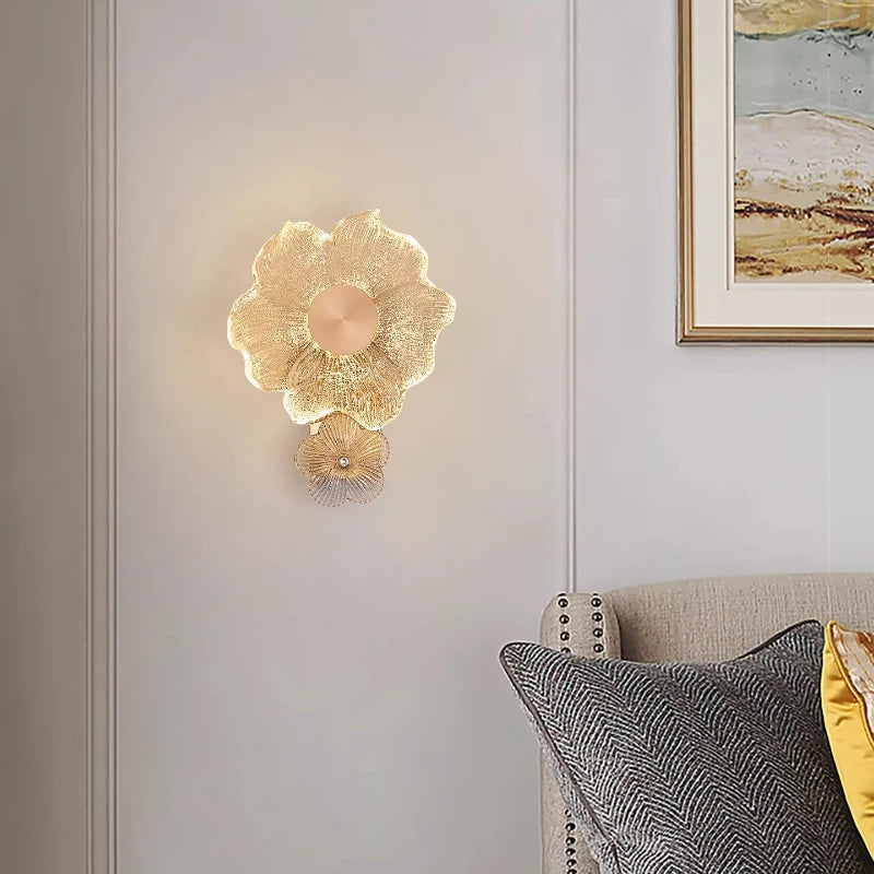 A gold flower glass wall sconce installed on a paneled wall in a living area, with a sofa and pillows visible.