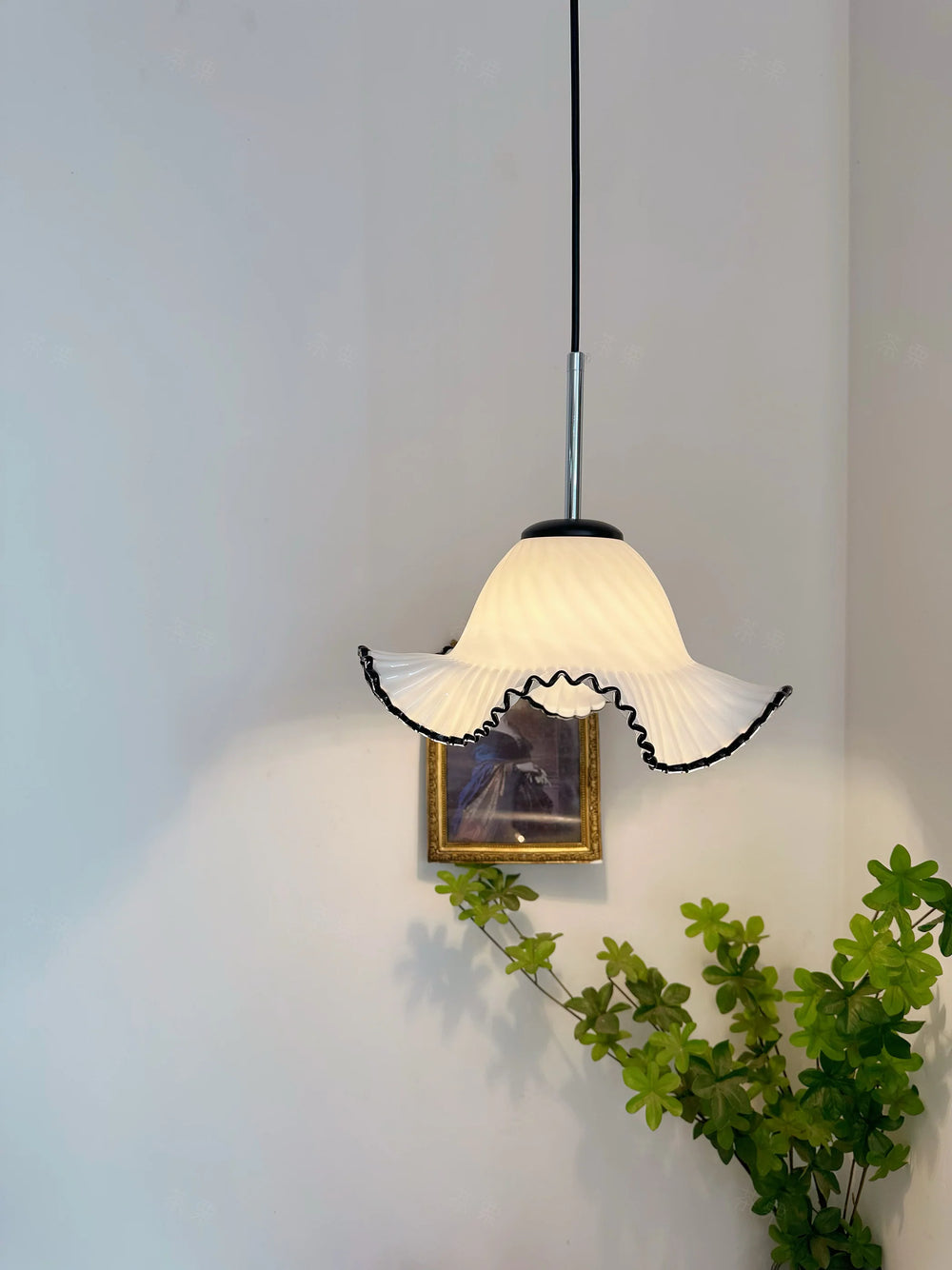 Pendant light with white lampshade and black trim hanging against a plain wall with a plant and framed picture.