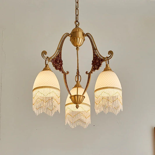 Front view of a polished brass chandelier with three white glass shades, each adorned with bead fringe, showcasing the detailed floral ceramic elements on its curved arms.