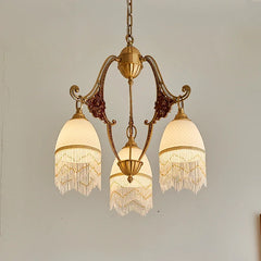 Front view of a polished brass chandelier with three white glass shades, each adorned with bead fringe, showcasing the detailed floral ceramic elements on its curved arms.