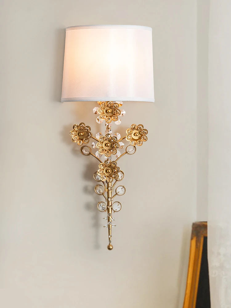 A gold floral crystal wall sconce with a white fabric shade mounted on a light-colored wall.