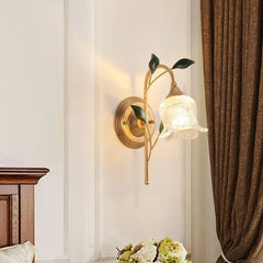Gold flower wall sconce with clear glass shade mounted on a white wall next to a wooden bed frame and a brown curtain.