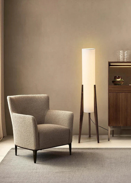 A minimalist floor lamp with a black walnut base and a tall, cylindrical white fabric shade, standing next to a textured armchair in a room with muted walls.