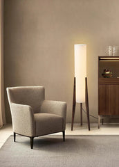 A minimalist floor lamp with a black walnut base and a tall, cylindrical white fabric shade, standing next to a textured armchair in a room with muted walls.