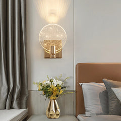 A gold and clear bubble crystal wall sconce installed in a bedroom, positioned above a bedside table with a geometric vase and flowers.