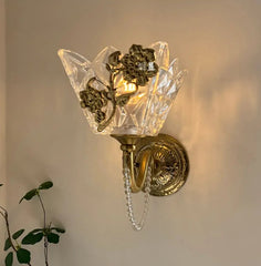 Side view of a gold wall sconce with a clear glass shade and floral detail, showing the curved arm and a hanging crystal chain detail.