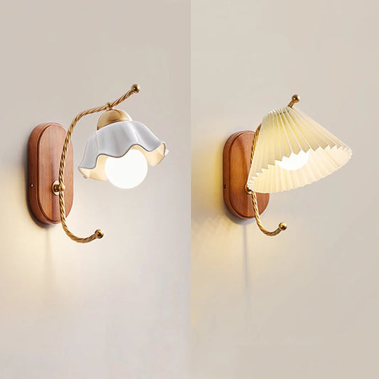 Two wall sconces side-by-side, one with a white ceramic flower shade and the other with a pleated cream fabric shade, both mounted on wooden bases with gold arms.