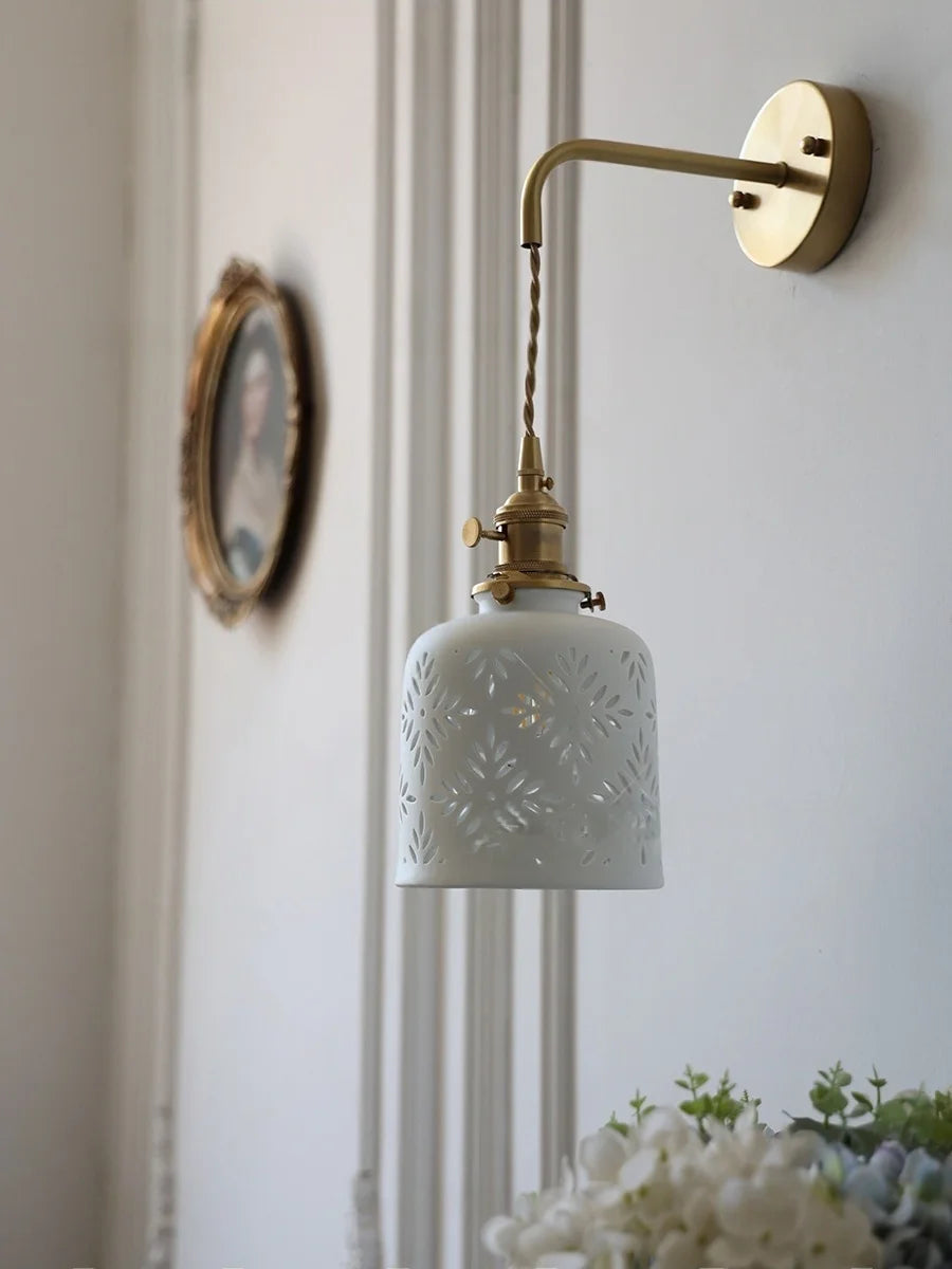 Wall-mounted lamp with decorative ceramic shade and brass arm on a light-colored wall.