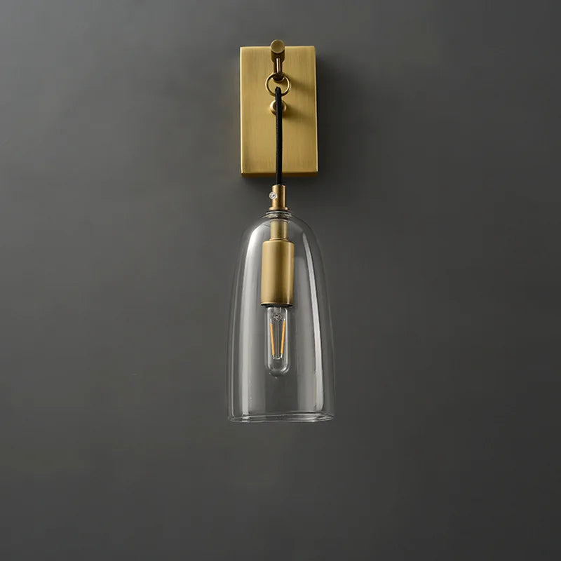 Close-up of a gold and clear glass bell wall sconce on a dark gray wall, showcasing the glass shade and bulb.
