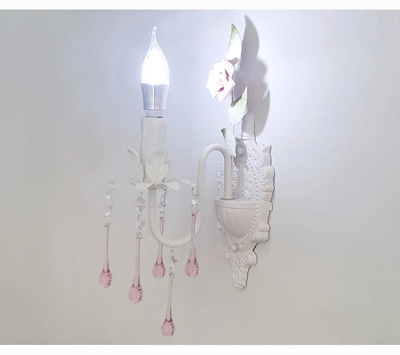 View of a white wall sconce with pink crystal droplets, the light illuminating the decorative rose accent on the backplate.