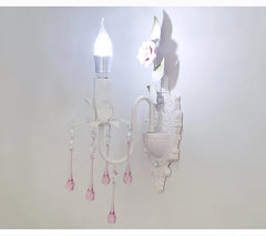 View of a white wall sconce with pink crystal droplets, the light illuminating the decorative rose accent on the backplate.