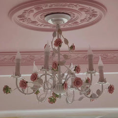 White iron chandelier with pink roses and green leaves, installed on a pink ceiling with decorative molding.