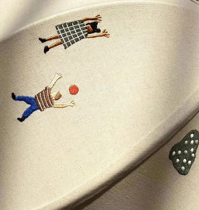 Embroidered design on a beige fabric with two figures and a ball.