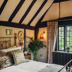 A wall-mounted brass sconce with a bell-shaped glass shade and tassel trim, illuminating a cozy bedroom with a canopy bed and dark wooden beams.