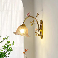 A gold floral wall sconce with a fluted glass shade is illuminated against a white wall, with green foliage visible in the foreground and a sheer curtained window in the background.