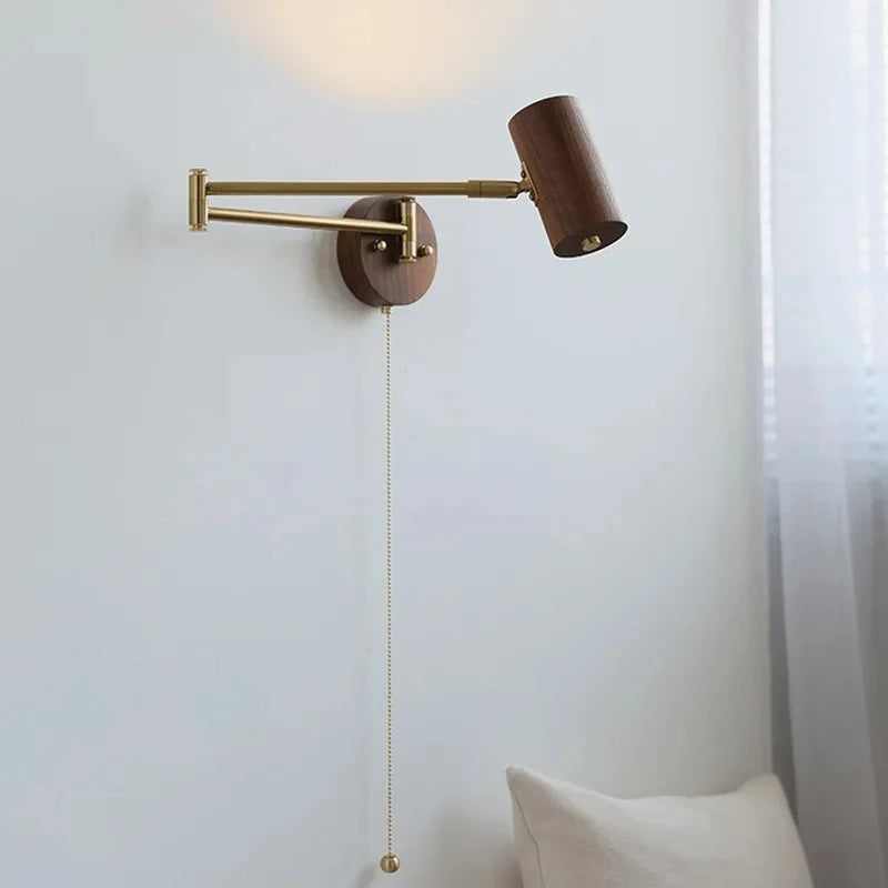 A wall sconce with a dark wood cylinder lamp head and a brass swing arm, with a brass pull chain visible, casting a warm light on the white wall.