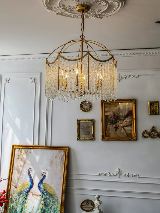 Gold crystal chandelier with cascading droplets installed in a decorated white room with framed artwork on the walls.