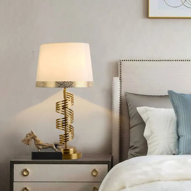 A gold spiral table lamp with a white conical fabric shade on a nightstand beside a bed and pillows.