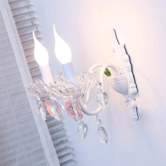 Side profile of a white wall sconce with two flame-tip bulbs, featuring pink ceramic roses and crystal accents, mounted on a white wall.