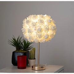 A white floral rose bud table lamp with a gold base and stem, placed on a nightstand next to potted plants.