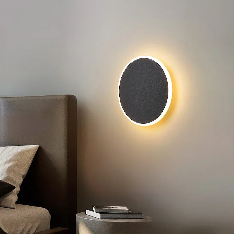 A single black circular wall sconce with a warm halo effect mounted on a grey wall next to a bed.