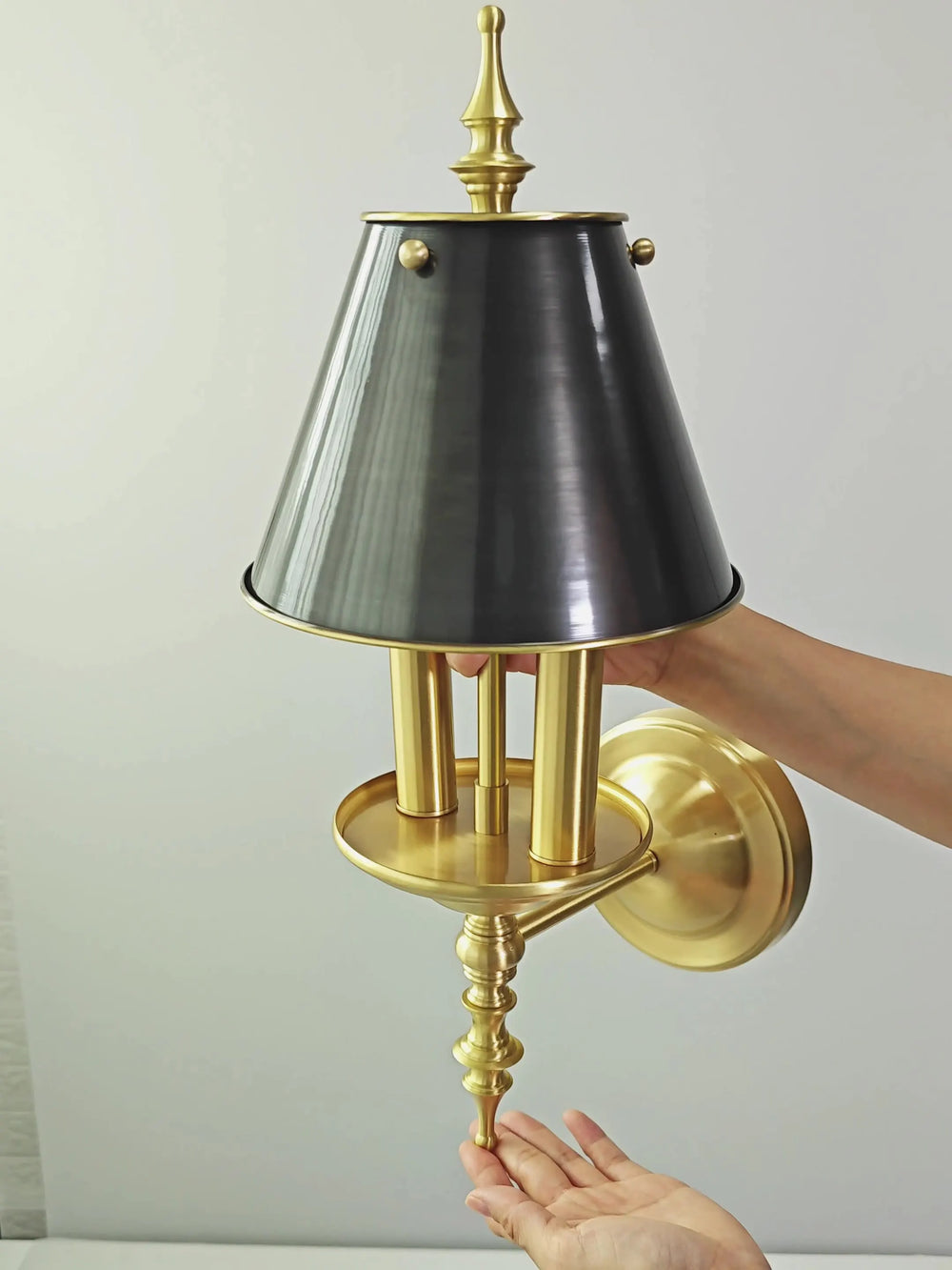 Wall-mounted lamp with black shade and gold base held by a hand against a light gray background