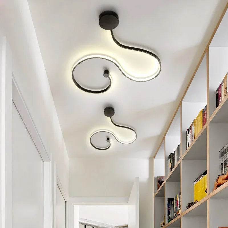 Two black wave-shaped LED ceiling lights with warm light mounted on a white ceiling in a hallway with bookshelves.