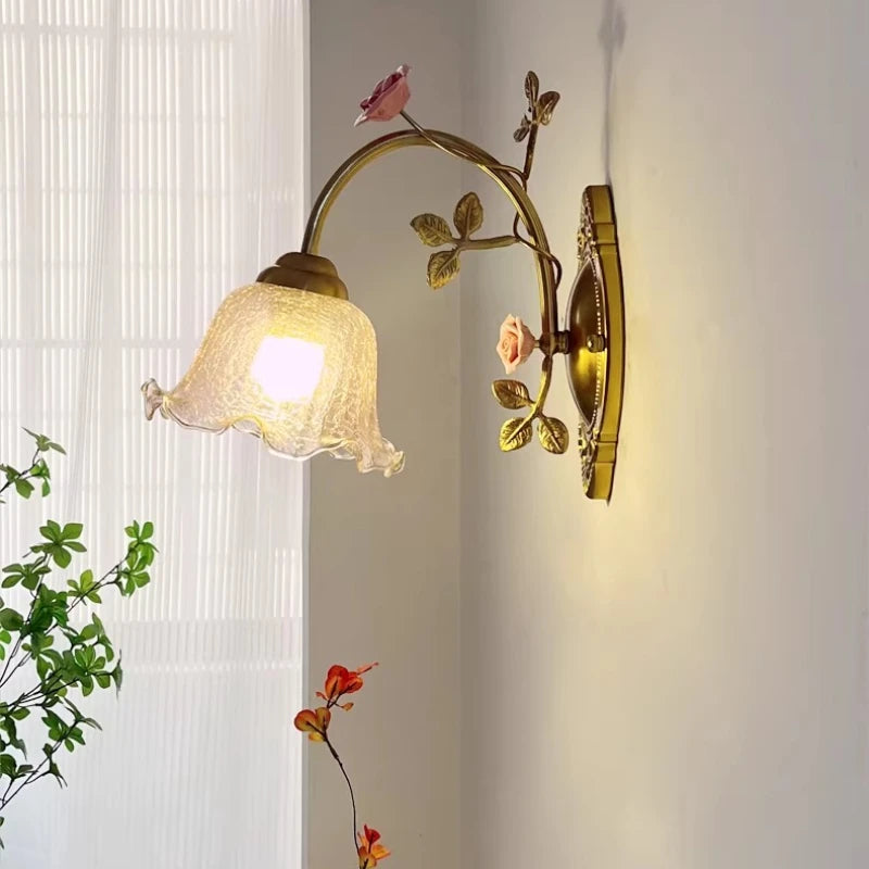 Side profile of a gold floral wall sconce with a glass shade, showcasing its curved arm and leaf and rose embellishments against a white wall.
