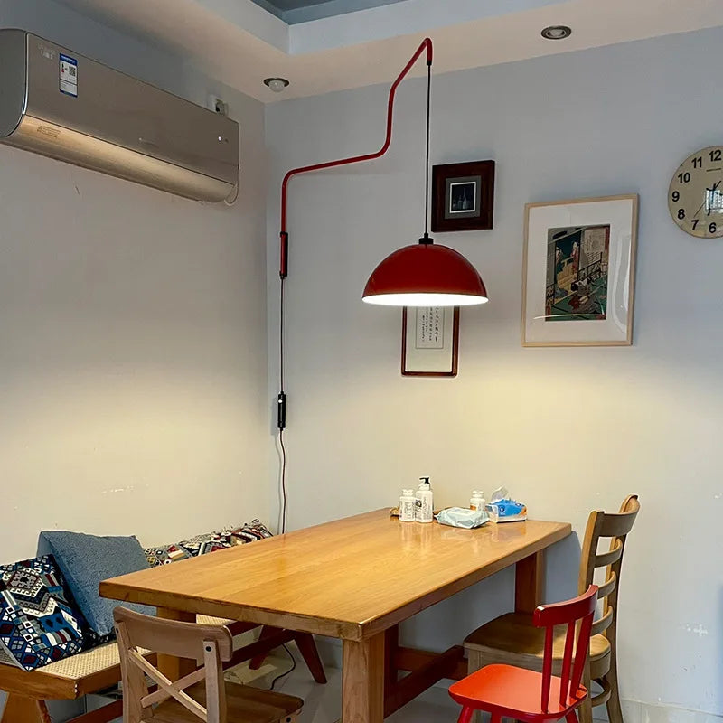 Red folding retractable wall lamp with a dome shade above a wooden dining table, near artwork on the wall and an air conditioning unit.