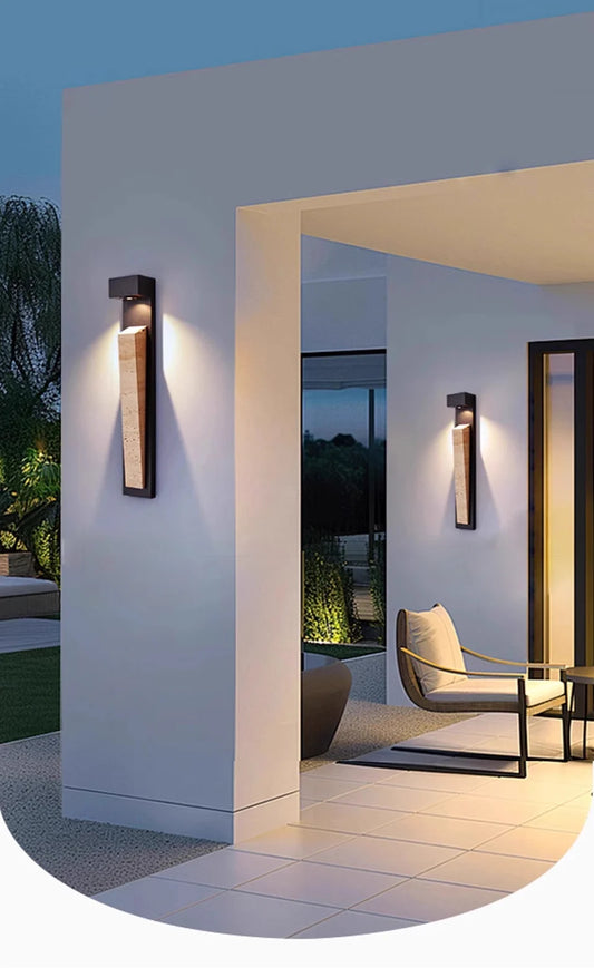 A modern black wall sconce with a natural stone insert mounted on a white outdoor wall, illuminated at dusk.