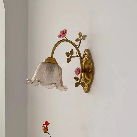Close-up view of a gold floral wall sconce featuring a textured glass shade and pink rose accents, mounted on a white wall.
